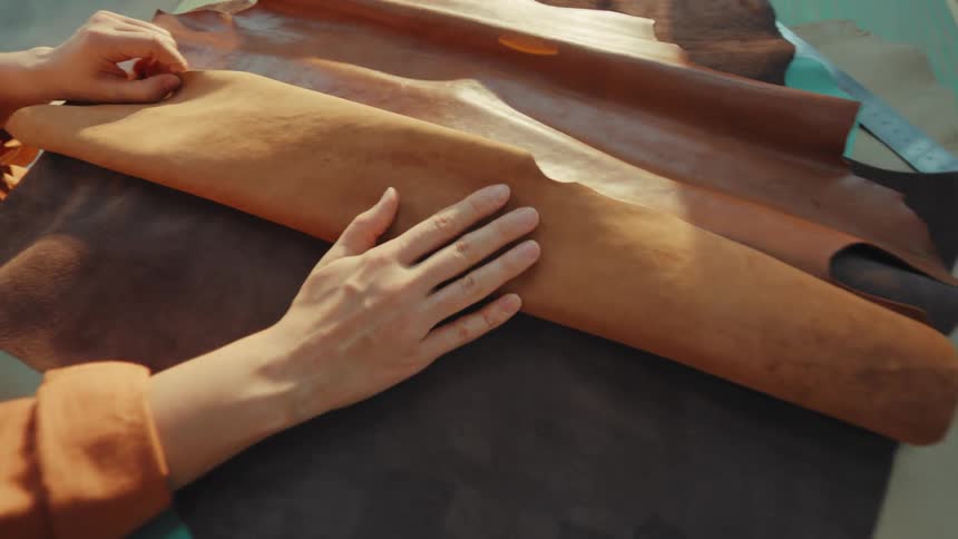 A female artisan carefully examines different pieces of natural leather, feeling the texture with her hands in a sunlit workshop