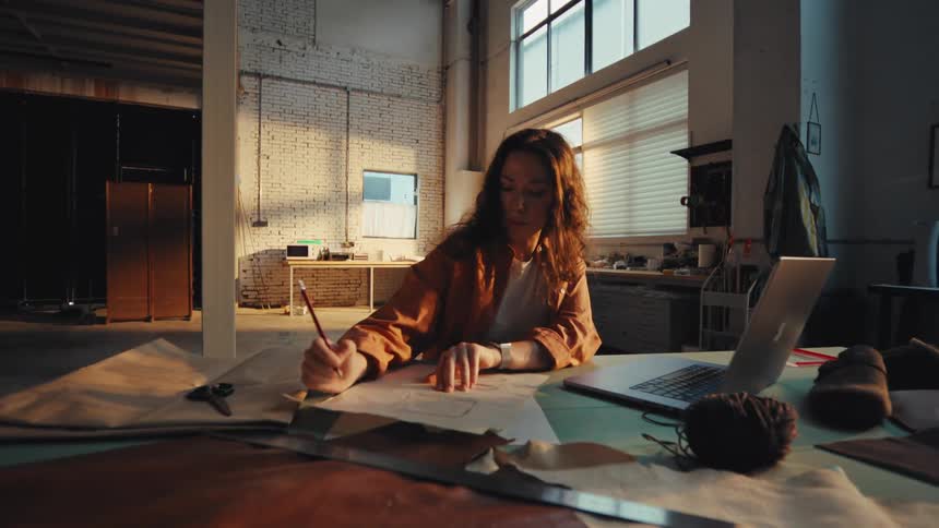 A focused female artisan sketches a design pattern on paper at a large workbench in her sunlit industrial workshop