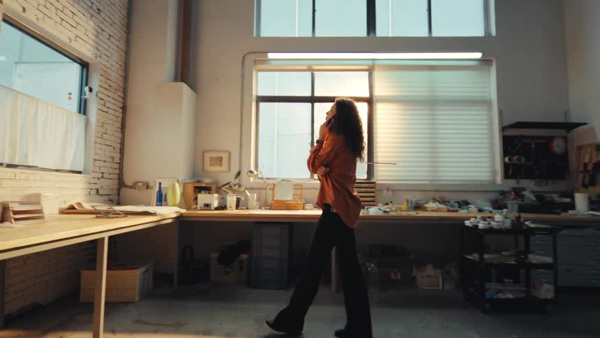 A young woman with curly hair talks on the phone while slowly pacing around her sunlit industrial workshop in slow motion