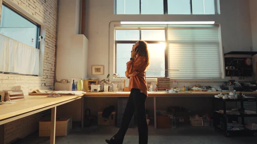 A young woman with curly hair talks on the phone while slowly pacing around her sunlit industrial workshop in slow motion