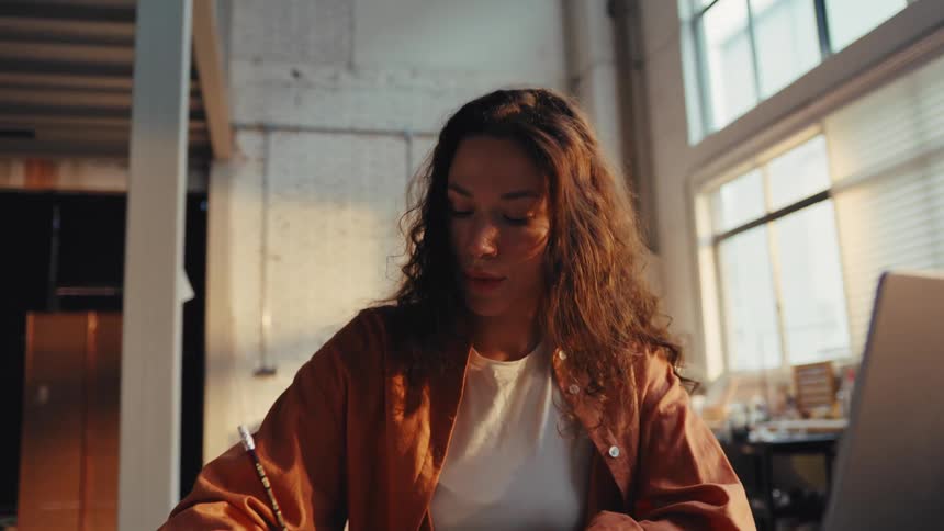 A focused female creative professional reviews a drawing and makes notes while working on her laptop in a sunlit industrial loft