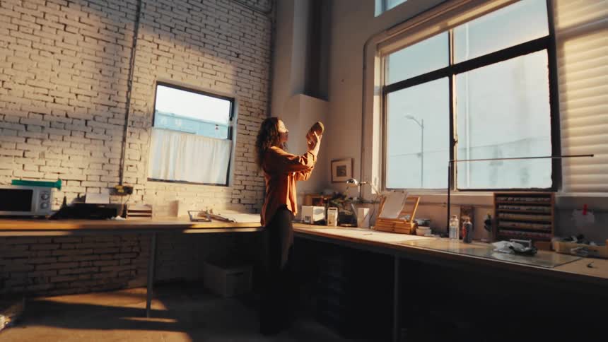 A young female creator carefully inspects her custom shoe design by the window in her sunlit, industrial studio