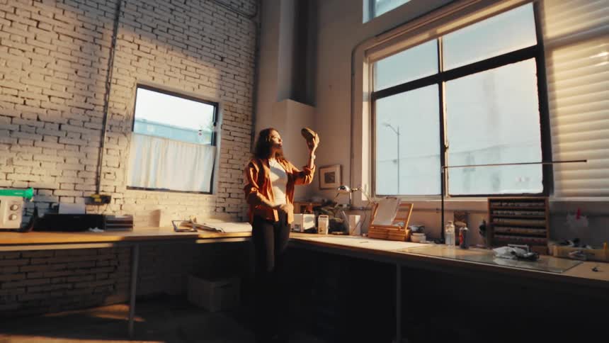 A young female creator carefully inspects her custom shoe design by the window in her sunlit, industrial studio