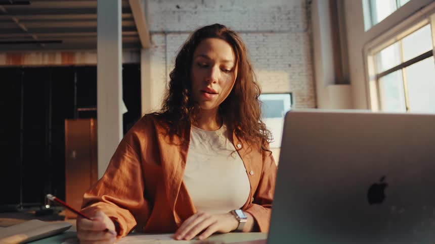 A creative young woman sketches a new design at her desk while working on her laptop in a warm, sunlit industrial loft studio