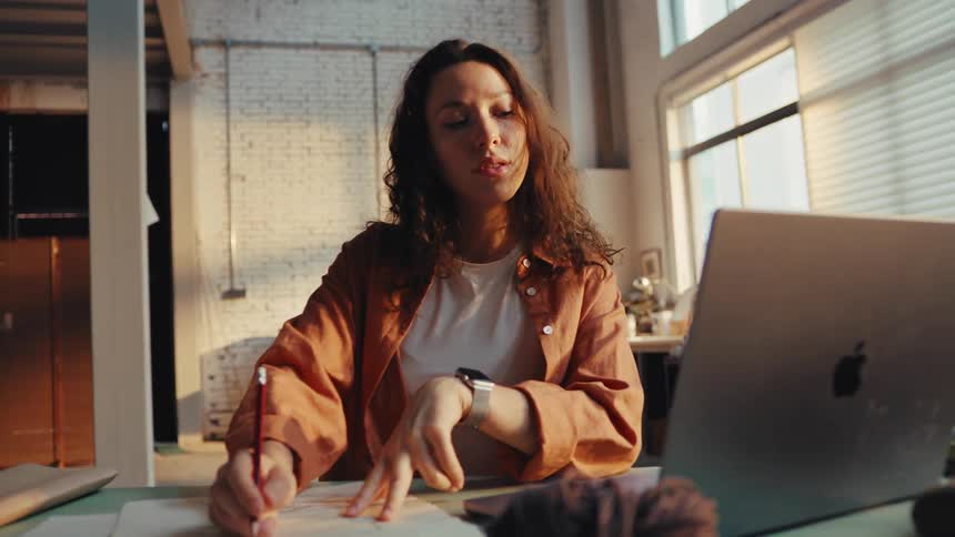 A creative young woman sketches a new design at her desk while working on her laptop in a warm, sunlit industrial loft studio