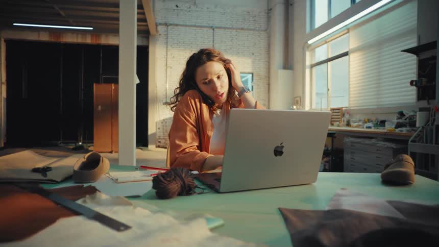 Female footwear designer multitasks on a phone call while using her laptop and sketching new designs in her creative workshop