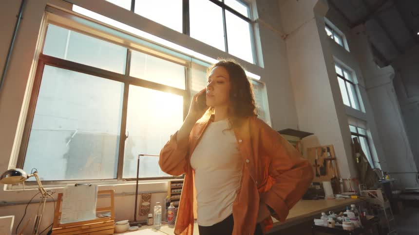 A young woman stands by a large window in her sunlit studio workshop while talking on her phone