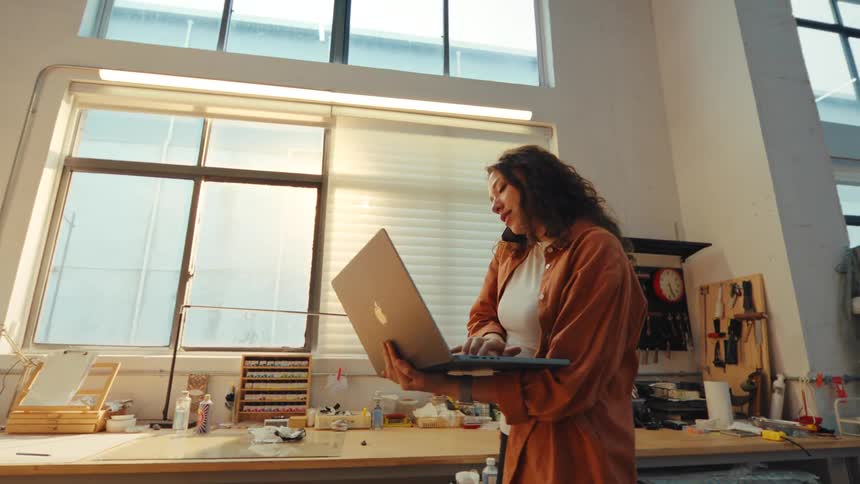 A young female artist multitasks in her sunlit studio, talking on the phone while working on her laptop