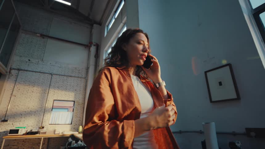 A low angle slow motion shot of a thoughtful young woman talking on her smartphone inside a sunlit industrial loft studio
