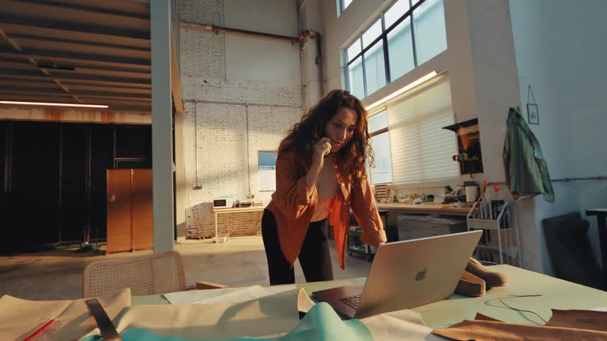 A young female artisan manages her business, talking on the phone and using a laptop in her sunlit industrial workshop