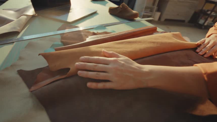 A female artisan gently unrolls and feels the texture of a piece of brown leather in her sunlit workshop
