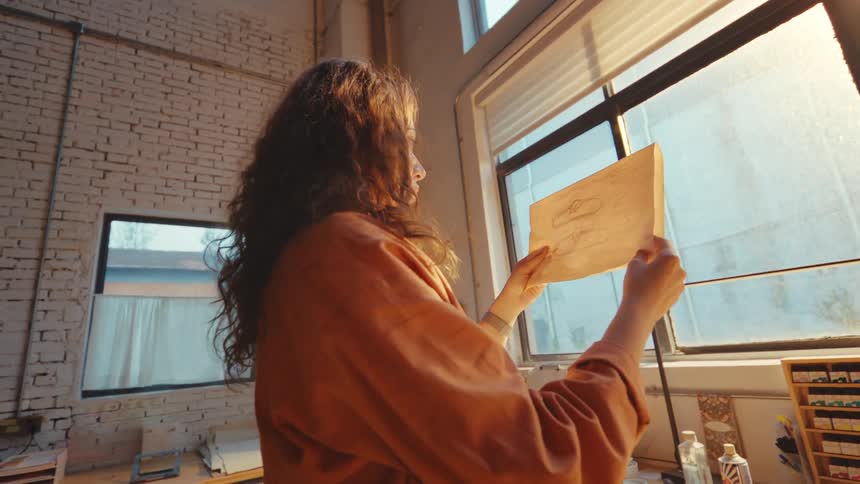 A female shoe designer examines her sketch against the warm light of a window in her art studio