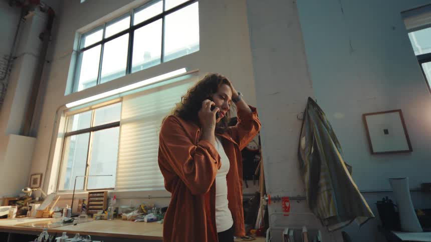 A young female artist has a dynamic phone conversation in her sunlit, industrial loft style art studio