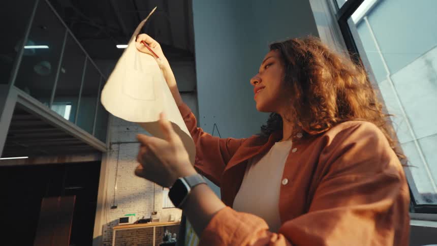 A pensive female artist evaluates her sketch in the warm, golden light of sunset streaming through her studio window