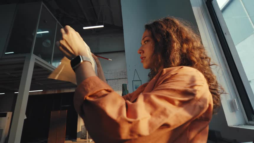A pensive female artist evaluates her sketch in the warm, golden light of sunset streaming through her studio window