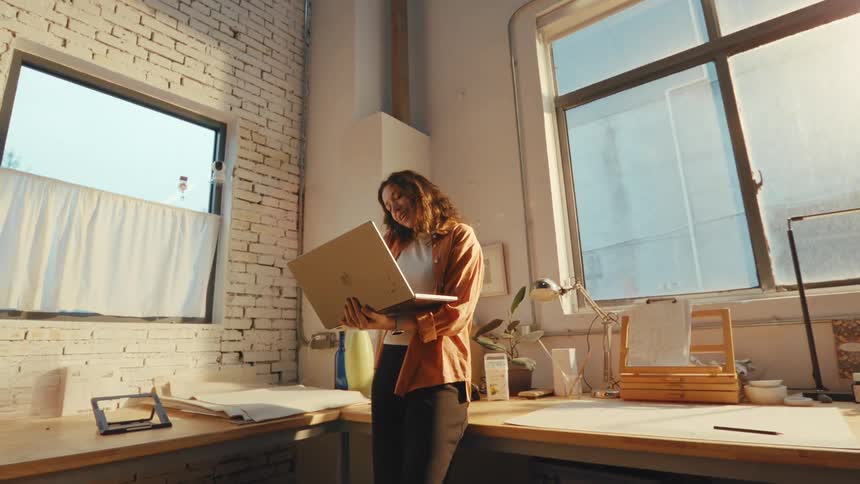 A cheerful female architect talks on the phone while holding her laptop in a sun-drenched industrial loft studio