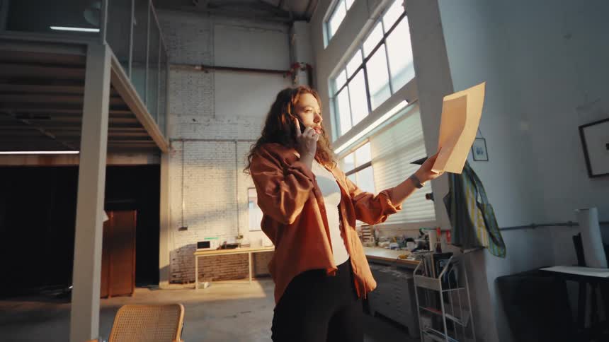 A female designer multitasks in her sunlit industrial workshop, sketching a design and taking a business call