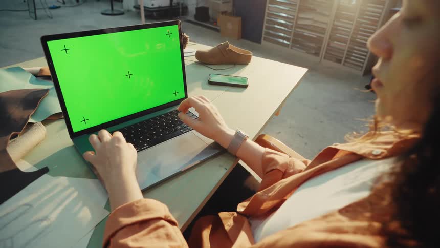 Over-the-shoulder shot of a female artisan working on a laptop with a green screen in her sunlit design studio