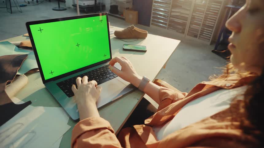 Over-the-shoulder shot of a female artisan working on a laptop with a green screen in her sunlit design studio