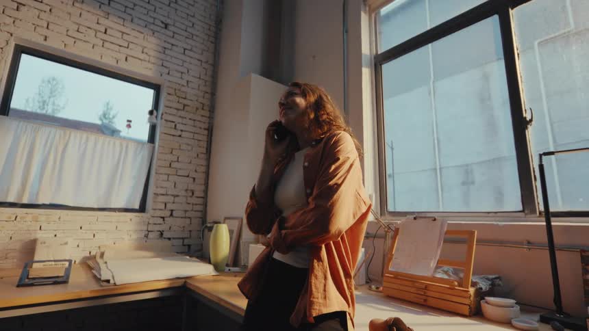 A happy woman smiles while enjoying a pleasant phone conversation in her sun-drenched industrial loft studio