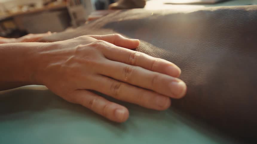 A craftsperson's hand gently glides over a piece of high-quality brown leather, feeling its texture in a sunlit workshop