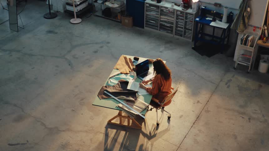 High-angle view of a female designer sketching at her drafting table, bathed in sunlight in her spacious industrial loft studio