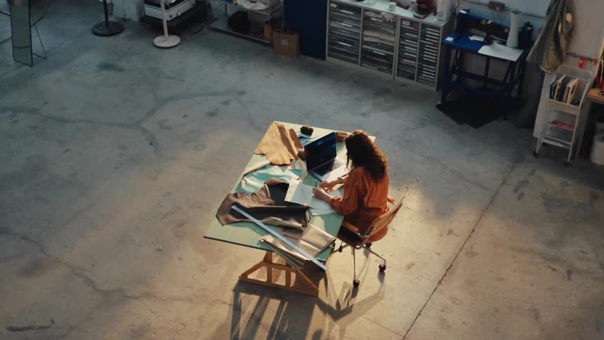 High-angle view of a female designer sketching at her drafting table, bathed in sunlight in her spacious industrial loft studio