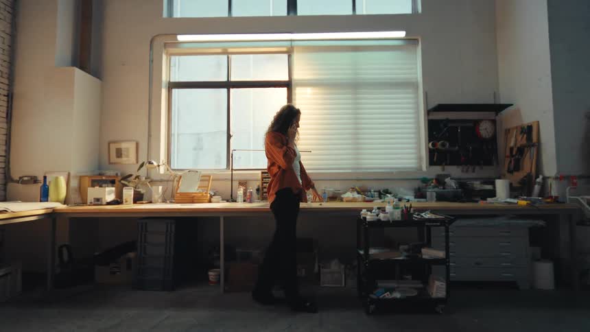 Slow motion shot of a young woman walking through her industrial studio while talking on the phone