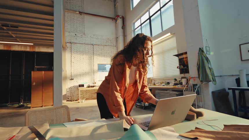A smiling young female designer works on a laptop and has a video call in her sun-drenched industrial craft studio