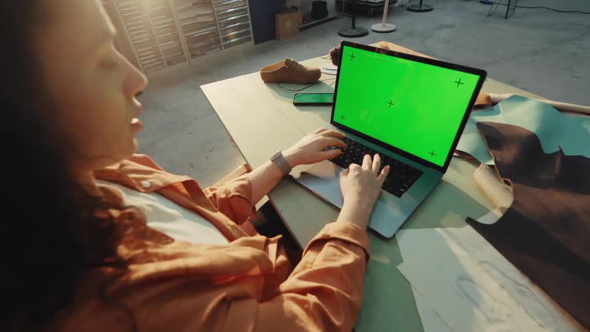 Over-the-shoulder view of a female shoemaker working on a laptop with a green screen in her sunlit artisan workshop