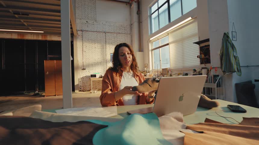 A female footwear designer has a video call on her laptop, showcasing a new shoe design from her sunny workshop