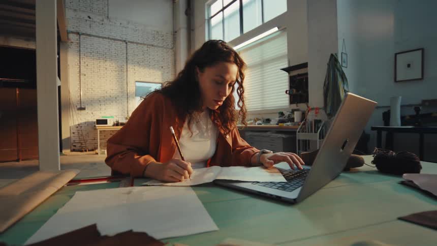 A creative female artisan diligently works on her laptop and sketches a new design pattern on paper in her spacious, sun-drenched workshop