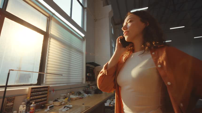 A young female artist stands in her sun-drenched industrial studio, having a thoughtful conversation on her smartphone