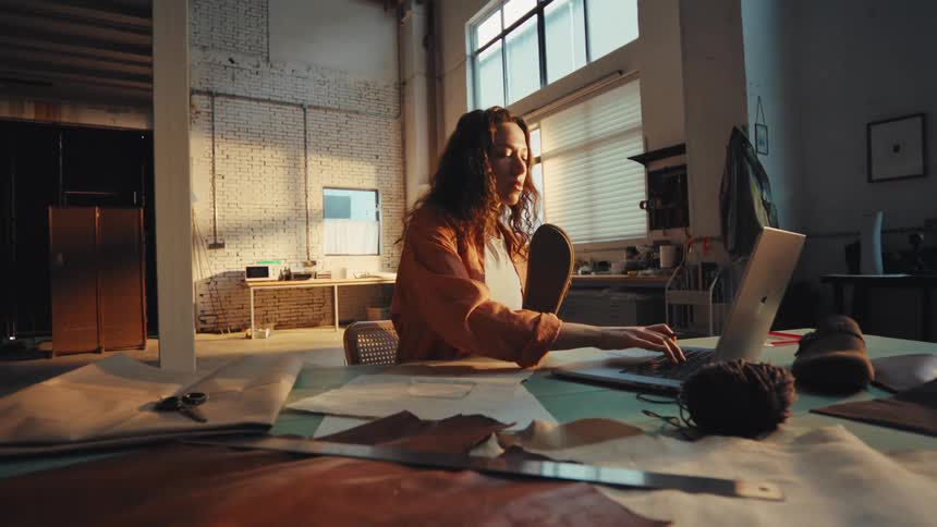 A female footwear designer sketches a new shoe model while examining a sole and using her laptop in a sunlit industrial workshop