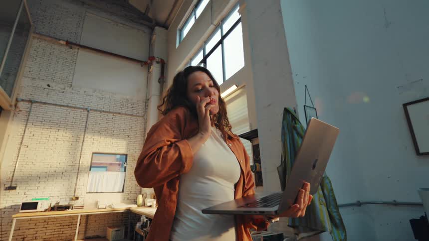 A busy female entrepreneur smiles while multitasking on a phone call and her laptop in a sunlit industrial workshop