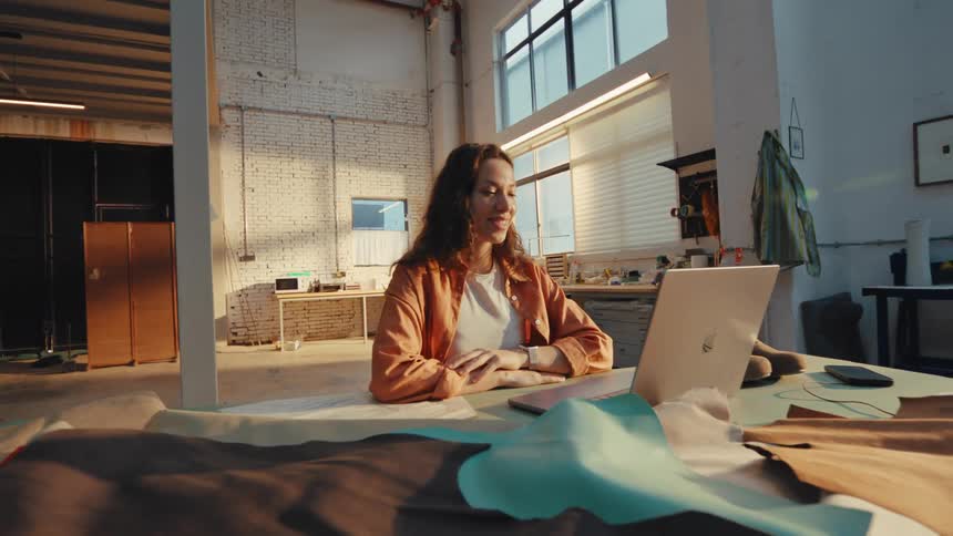 A young female leather artisan has a video call on her laptop while sitting at a workbench in her sunlit studio