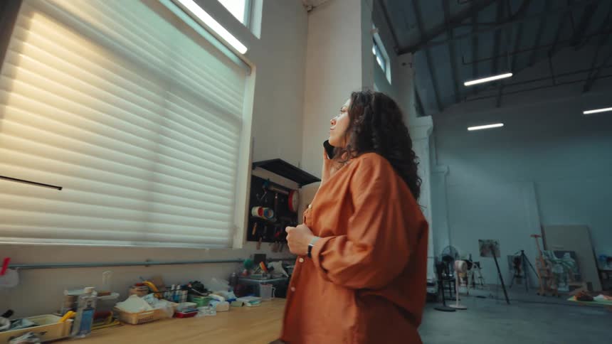 A young female artist with curly hair talks on the phone in her large, sunlit studio