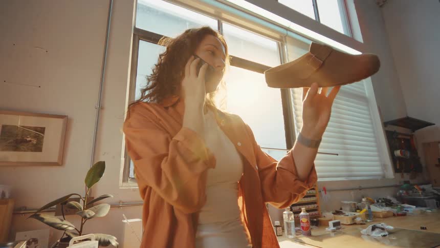 A female shoe designer inspects a handmade suede clog while talking on the phone in her sun-drenched workshop