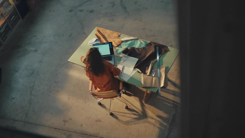 High-angle view of a female fashion designer working on her laptop and sketching patterns in a sunlit industrial studio