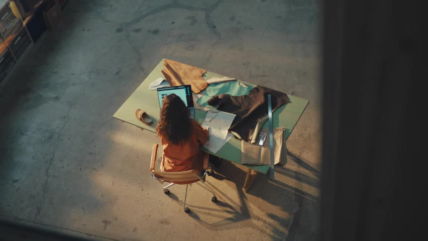 High-angle view of a female fashion designer working on her laptop and sketching patterns in a sunlit industrial studio