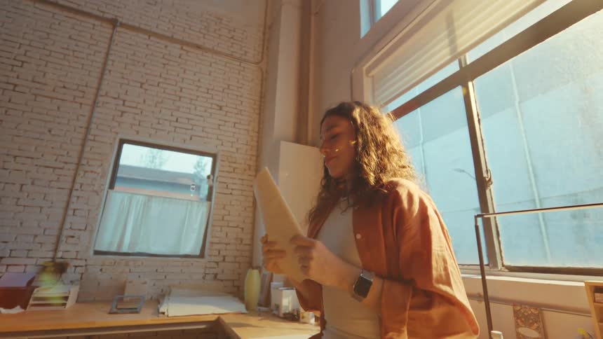 An inspired female artist with curly hair smiles as she examines her large sketch in a sun-drenched industrial studio