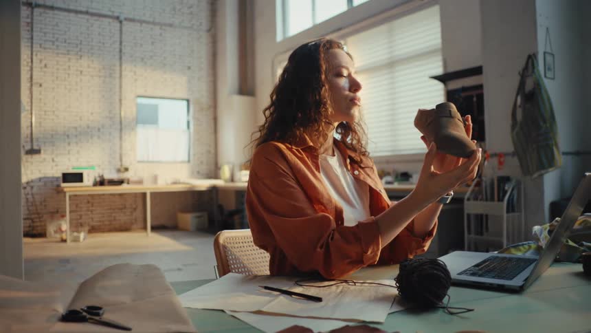 A young female shoemaker carefully inspects a newly crafted leather shoe in her sunlit industrial workshop