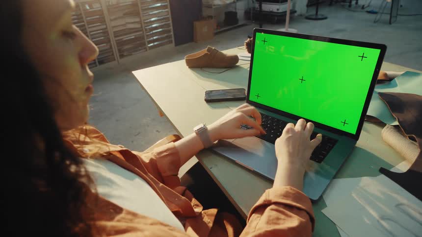 Over-the-shoulder view of a female artisan working on a laptop and holding a smartphone, both with green screens, in her sunlit workshop