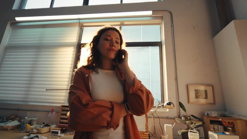 A young artist stands in her sun-drenched studio while listening to a serious phone call