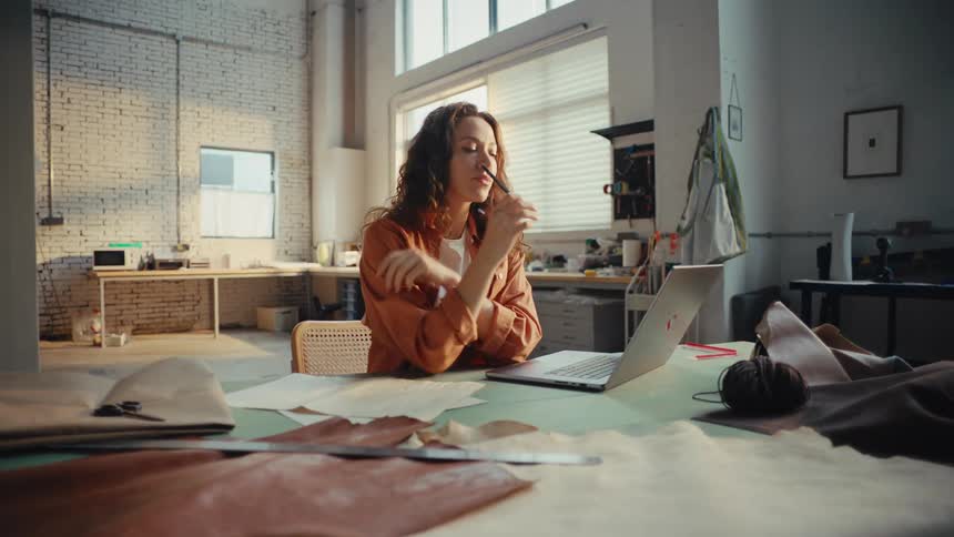 A creative young woman works as a designer in her studio, using a laptop for research and sketching ideas on paper amidst leather samples