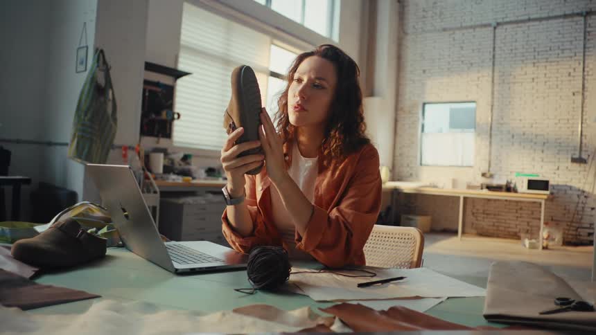 A thoughtful female shoemaker meticulously examines the sole of a handmade shoe in her sunlit artisanal workshop