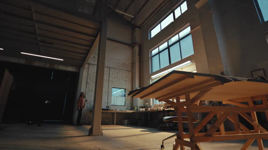 Female designer enters her large, industrial studio at dusk, turning on the lights and settling in at her workbench for a creative session