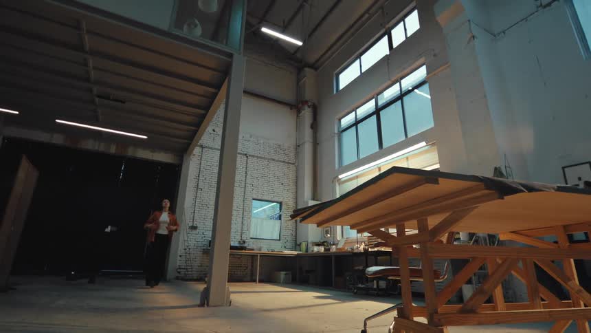 Female designer enters her large, industrial studio at dusk, turning on the lights and settling in at her workbench for a creative session