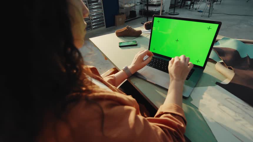 Over-the-shoulder shot of a female shoemaker working on a laptop with a green screen in her workshop
