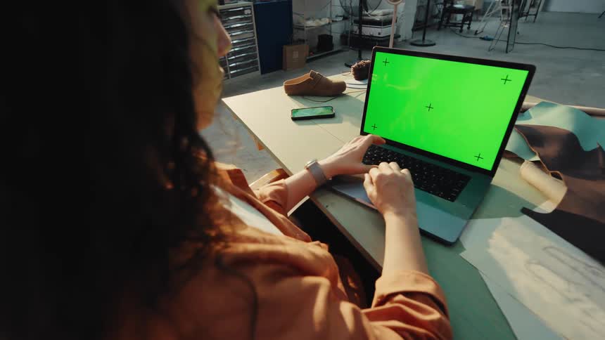 Over-the-shoulder shot of a female shoemaker working on a laptop with a green screen in her workshop